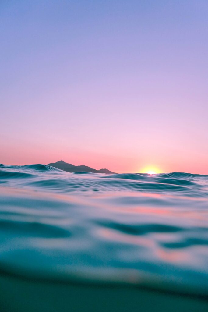 Beautiful zoomed-in photo of a sunset over the open ocean, where the water glistens in shades of blue under an orange sun. The sky above is tinted with a purple hue, adding a serene and vibrant backdrop.