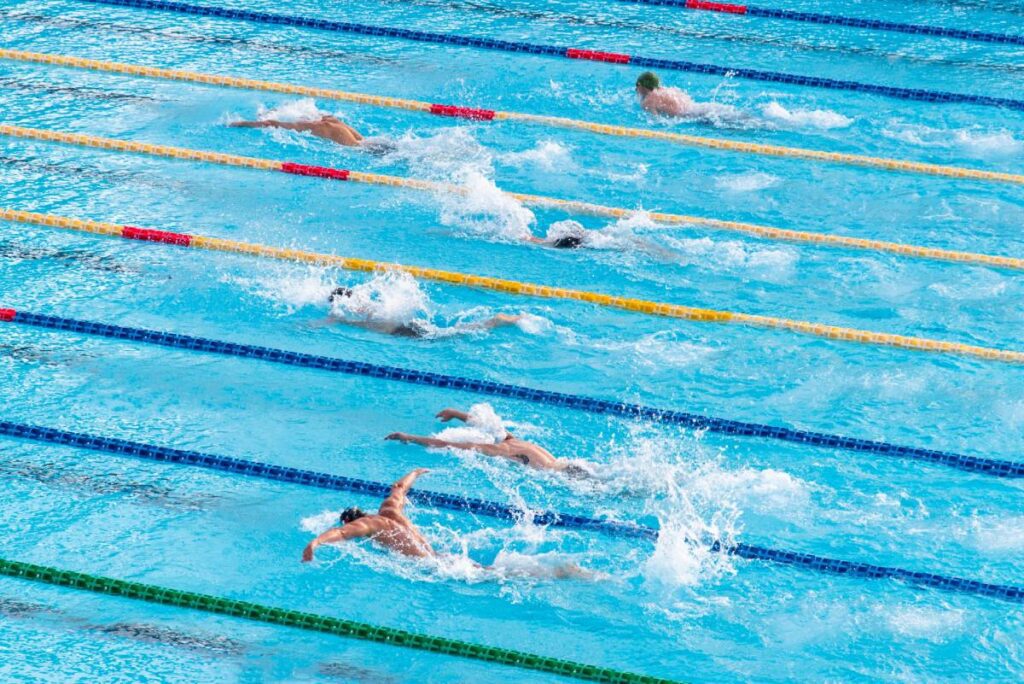 High view of a competitive swim race with multiple swimmers in various lanes performing the butterfly stroke. The pool's crystal blue water contrasts with the many colors of swim caps, creating an energizing and dynamic photo.