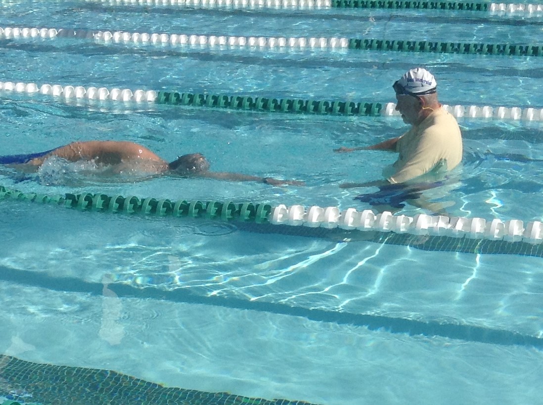 Coach David Guffey stands in a pool lane, wearing a white swim cap, as he coaches a swimmer on stroke techniques, demonstrating and giving feedback.