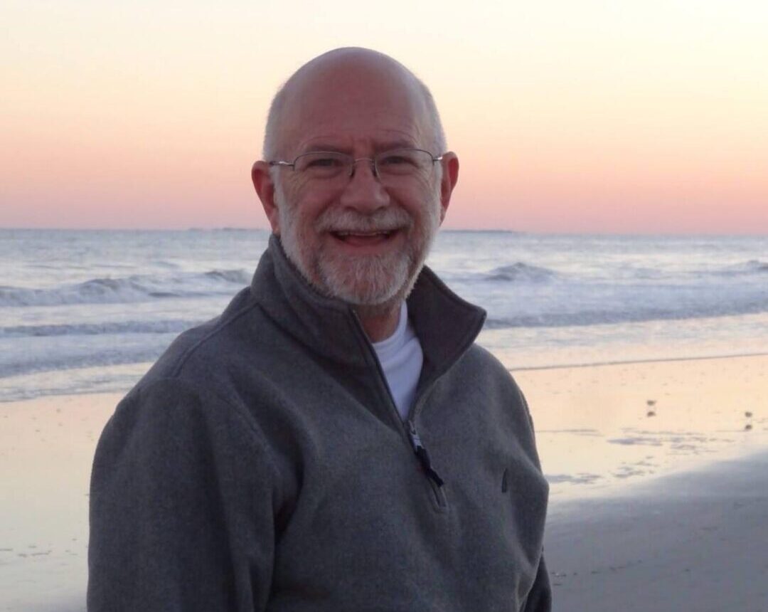 Coach David Guffey stands on the beach at sunset, cropped from the waist up, wearing a sweater pullover. He smiles with a very inviting demeanor, enhancing the warm and welcoming atmosphere of the scene.