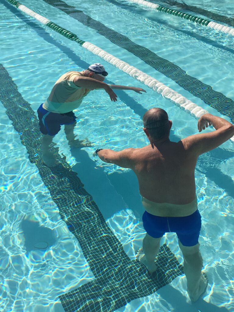 Coach David Guffey stands in a swim lane, demonstrating an arm movement to a student, emphasizing proper technique and form in a clear, instructive setting.