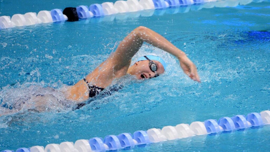 Female swimmer in full motion in a pool lane, performing a freestyle stroke during a competition. She is wearing a black bathing suit, swim cap, and goggles, showcasing her technique and speed.