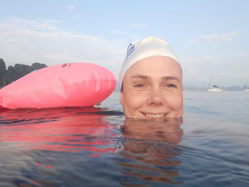 Female swimmer in open water, her head just breaking the surface enough to reveal a big smile. She wears a white swim cap and is accompanied by a pink flotation device, enhancing the joyful and buoyant atmosphere of the scene.