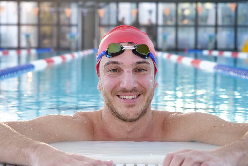 Male swimmer stands at the end of a swim lane, wearing a red swim cap and goggles on top of his head, smiling and feeling accomplished after his recent training session.