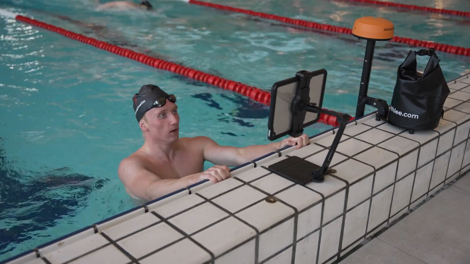 Male swimmer pauses at the end of the lane, wearing a black swim cap, to look at an iPad on a stand. He evaluates an Athlee video captured during his most recent lap, focusing on technique and performance improvements