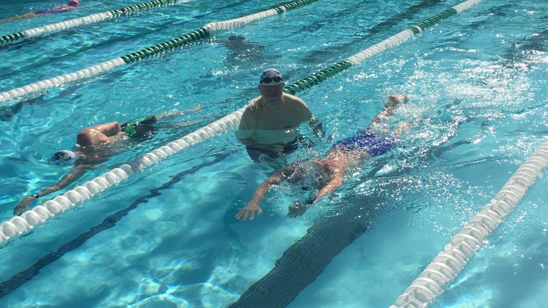 Coach David Guffey stands in a swim lane right beside a swimmer, closely analyzing and coaching the swimmer's stroke technique. Another swimmer is visible in the adjacent lane, adding a dynamic of shared training space.